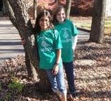 Two girls in green shirts standing next to a tree.